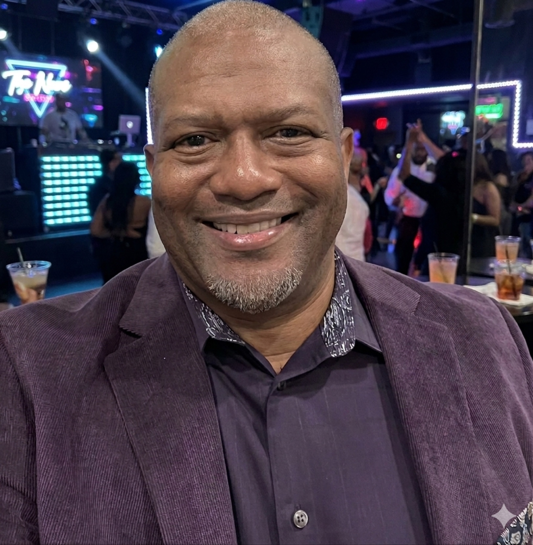 Smiling man in a purple blazer with a DJ booth and dance floor behind him