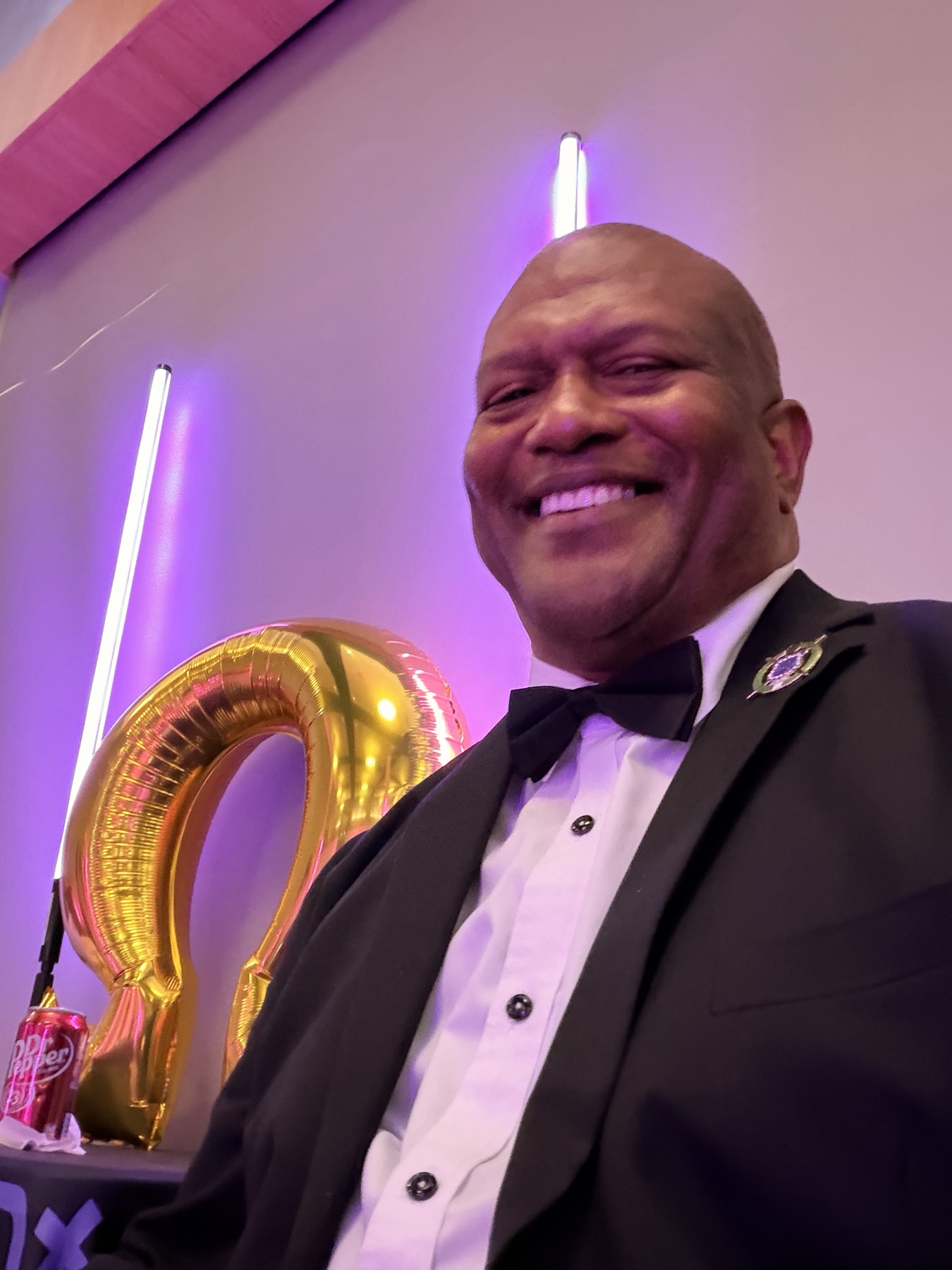 Man in a tuxedo smiling beside a gold Omega balloon and purple light