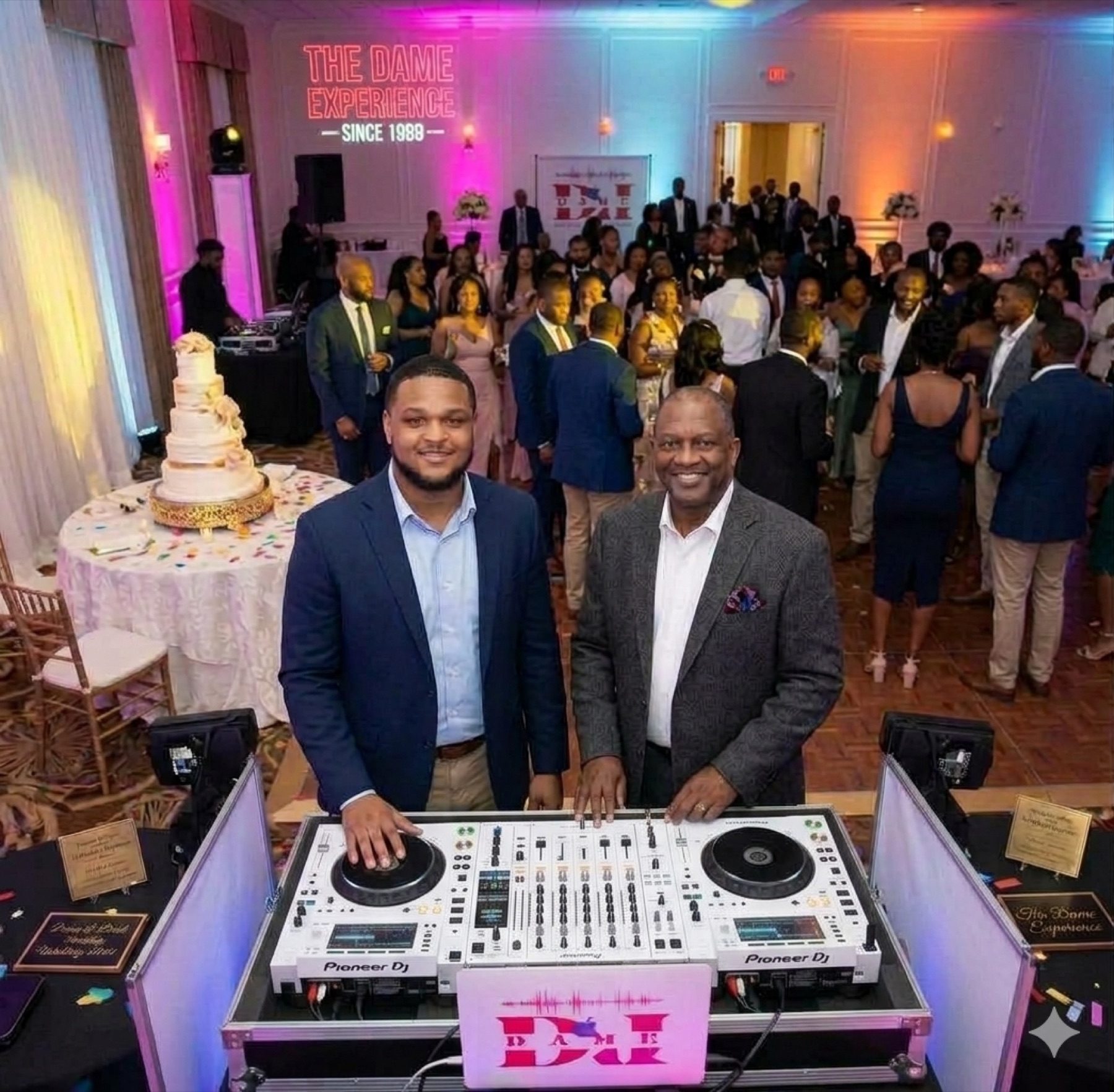 Two men standing behind a white Pioneer DJ controller at a reception