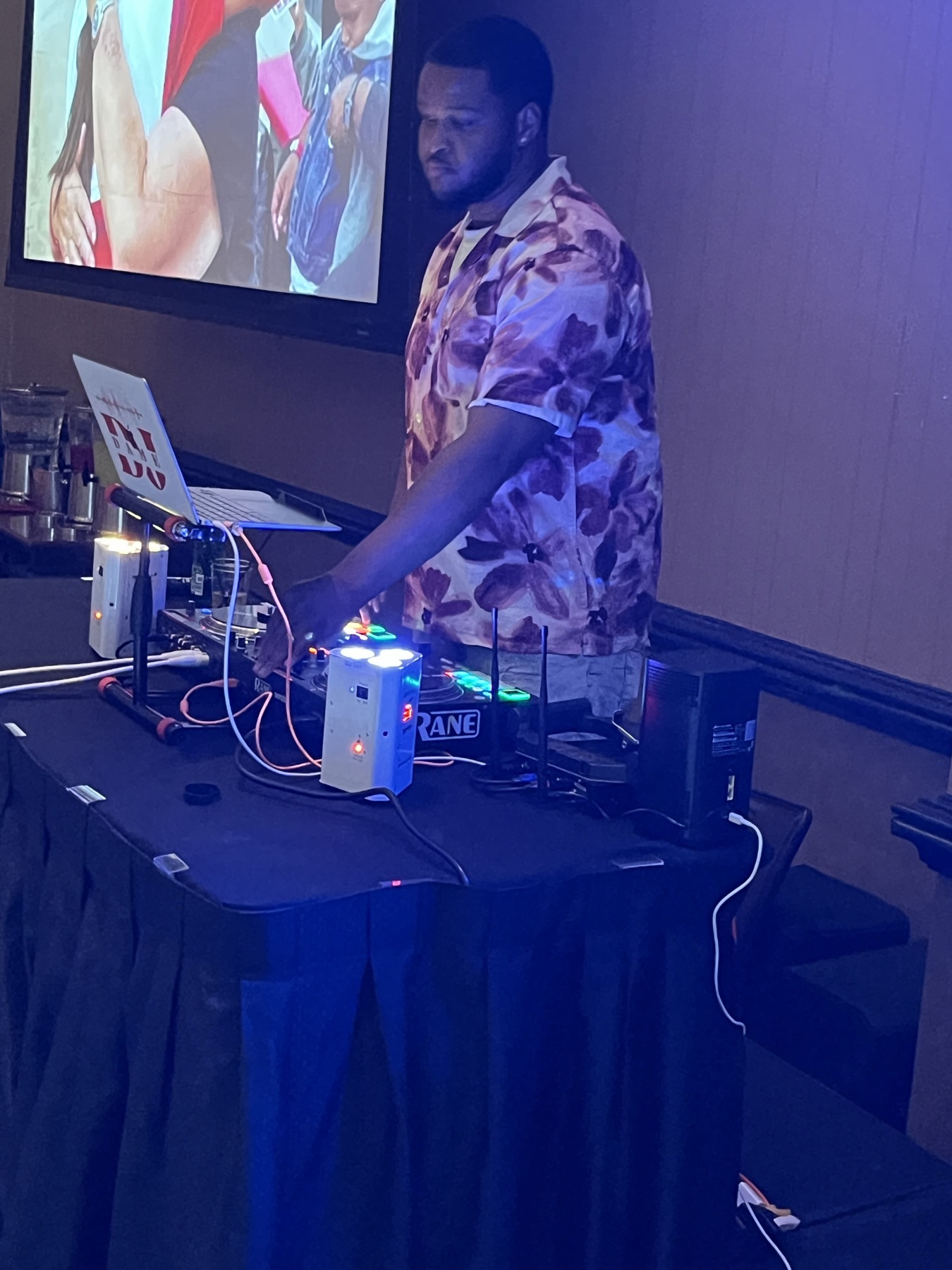 DJ in a floral shirt at a Rane controller with a projection screen behind