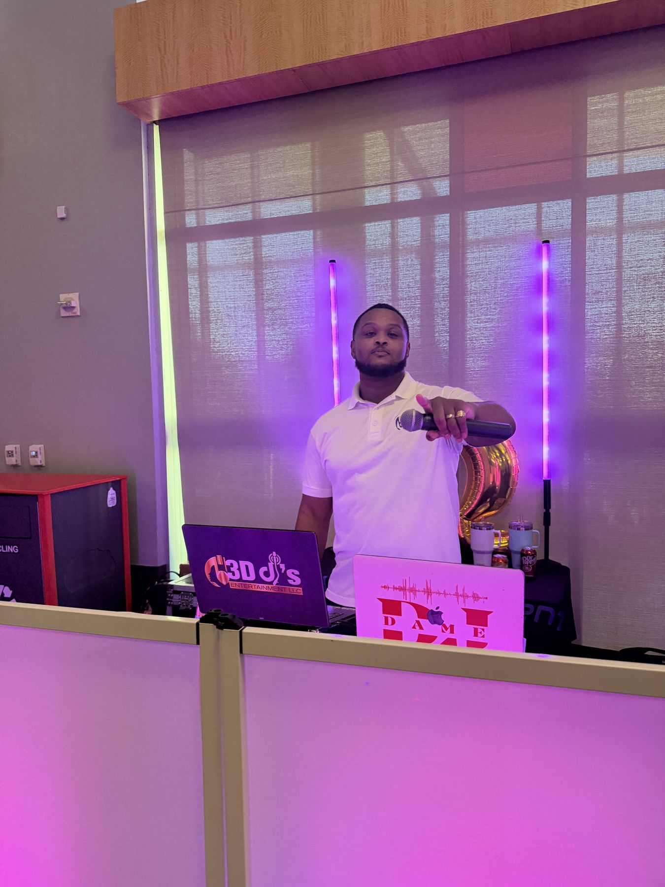 DJ holding a microphone behind laptops with 3D DJ's and DJ Dame logos