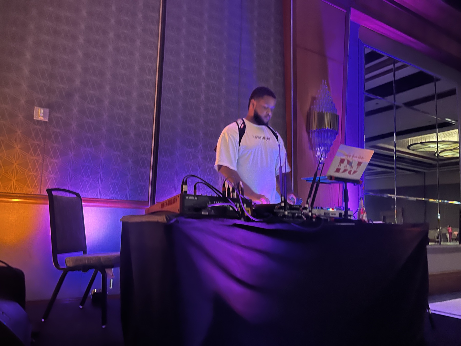 DJ at a draped table with a laptop and controller under purple uplighting