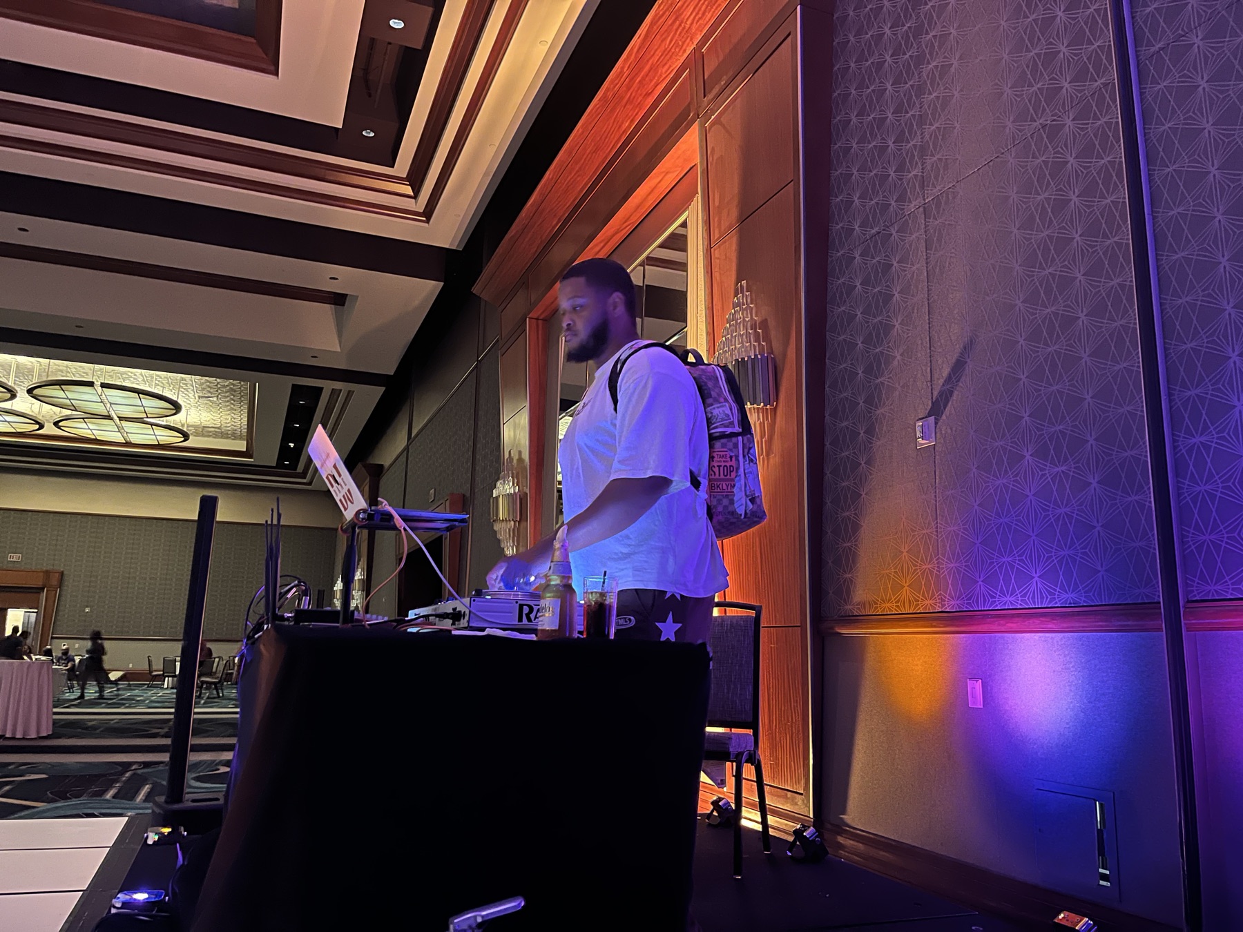 DJ wearing a backpack at a draped DJ table in a large ballroom