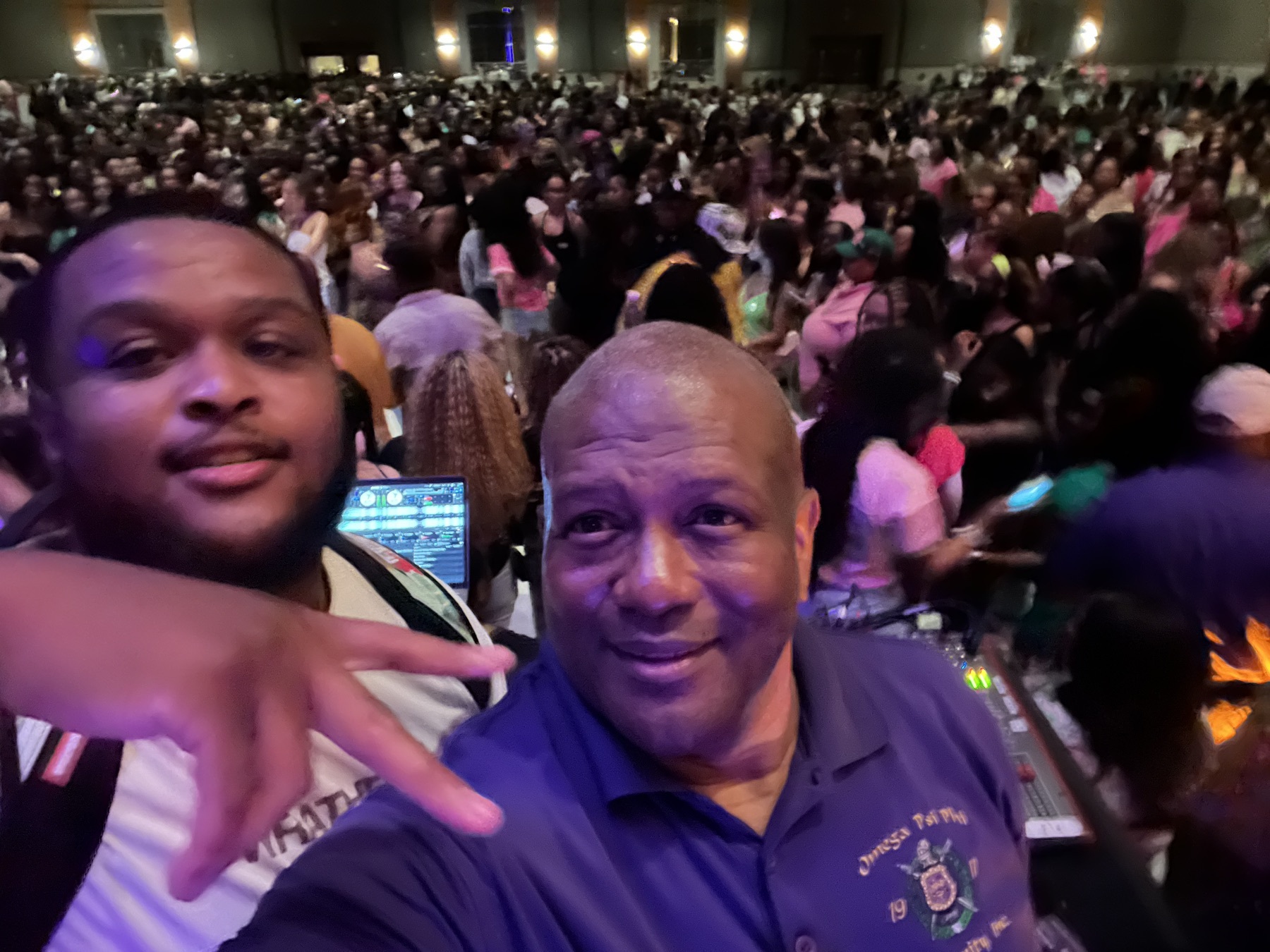 Two men taking a selfie in front of a large crowd near the DJ console
